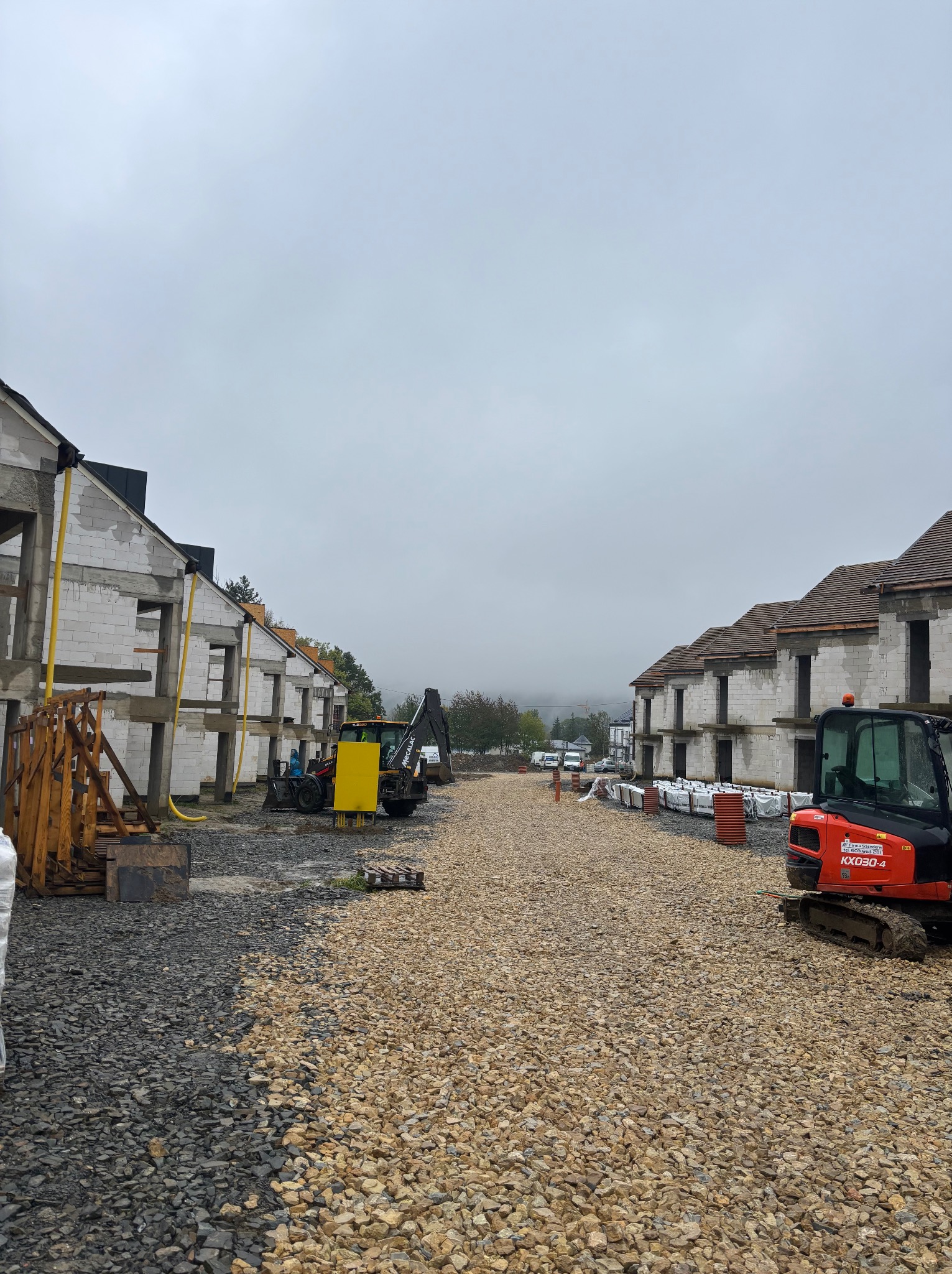 Budowa osiedla domów jednorodzinnych w trakcie realizacji. Widoczne surowe mury, maszyny budowlane i przygotowane podłoże. Pochmurne niebo w tle.