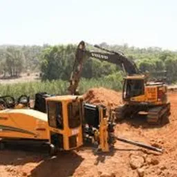 Żółta wiertnica pozioma sterowana obok koparki Volvo na placu budowy z wykopem, widoczne drzewa w tle.