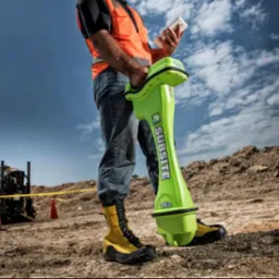 Pracownik w kamizelce odblaskowej obsługuje urządzenie Subsite TKa do lokalizacji podziemnych instalacji na placu budowy, w tle maszyna wiertnicza i pomarańczowe pachołki.