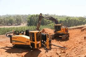 Żółta wiertnica pozioma sterowana obok koparki Volvo na placu budowy z wykopem, widoczne drzewa w tle.