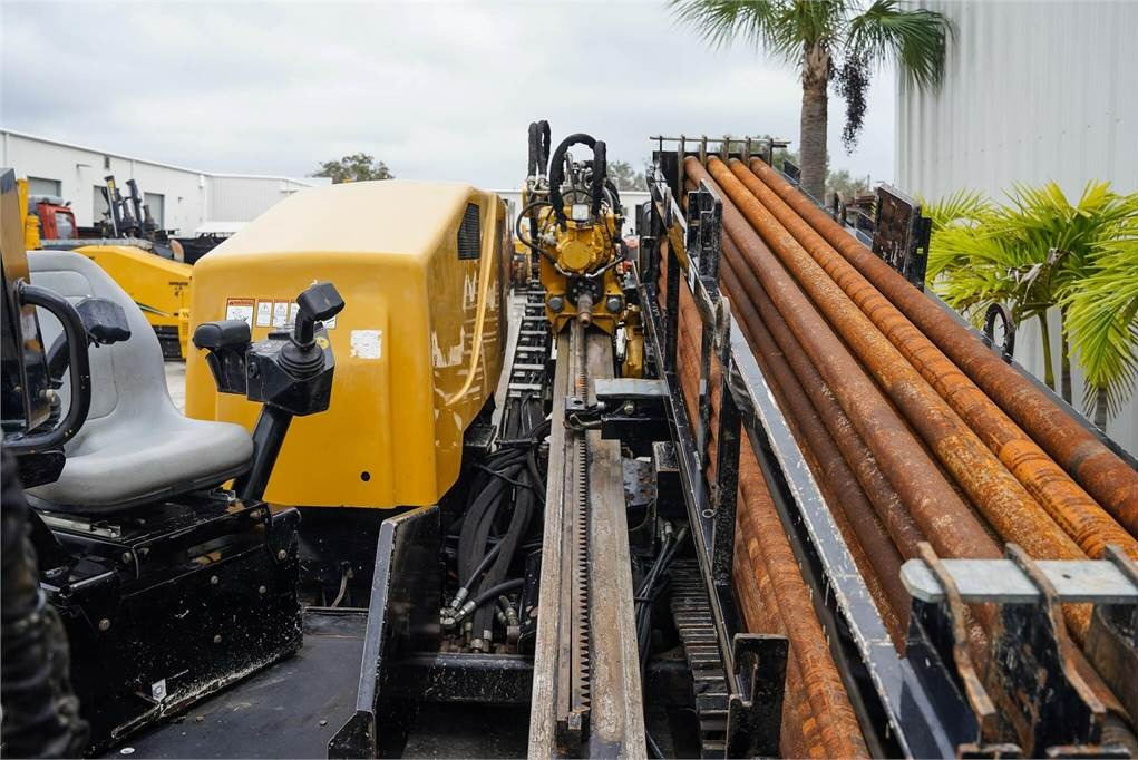 Żółta wiertnica kierunkowa z rzędem rdzawych rur na tle palm i budynku przemysłowego.