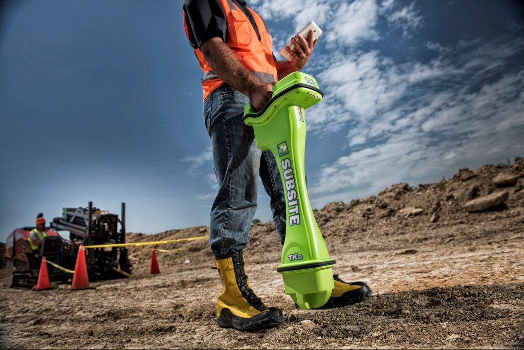 Pracownik w kamizelce odblaskowej obsługuje urządzenie Subsite TKa do lokalizacji podziemnych instalacji na placu budowy, w tle maszyna wiertnicza i pomarańczowe pachołki.