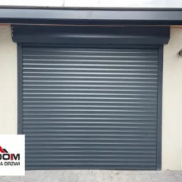 Zamknięta, szara brama rolowana z poziomymi panelami, zamontowana na jasnej elewacji budynku, widoczny fragment logo w lewym dolnym rogu.