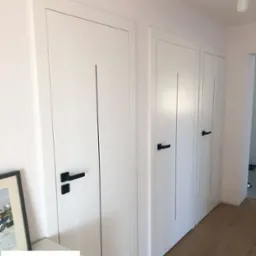 Trzy białe skrzydła drzwiowe z czarnymi klamkami i pionowymi frezowaniami, zamontowane w rzędzie w jasnym wnętrzu, widoczne z perspektywy.