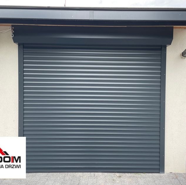 Zamknięta, szara brama rolowana z poziomymi panelami, zamontowana na jasnej elewacji budynku, widoczny fragment logo w lewym dolnym rogu.