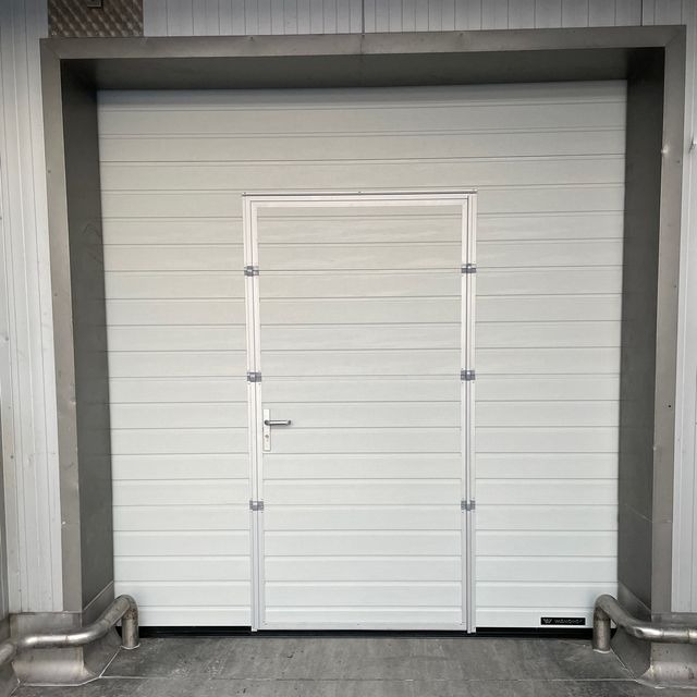 Zamknięta, segmentowa brama garażowa w kolorze białym z wbudowanymi drzwiami wejściowymi, widoczne metalowe ramy i zawiasy oraz rura odpływowa przy podłodze.