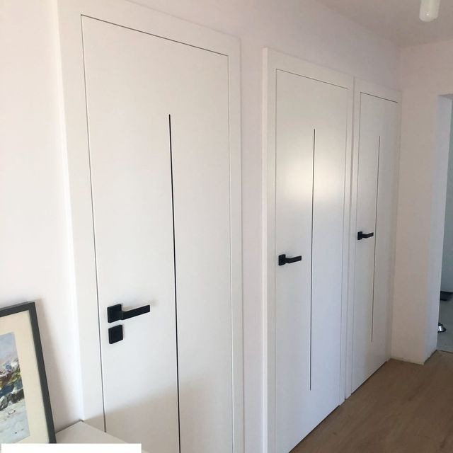 Trzy białe skrzydła drzwiowe z czarnymi klamkami i pionowymi frezowaniami, zamontowane w rzędzie w jasnym wnętrzu, widoczne z perspektywy.