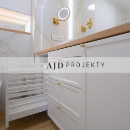 AJD PROJEKTY SPÓŁKA Z OGRANICZONĄ ODPOWIEDZIALNOŚCIĄ - Biała łazienka z marmurowymi ścianami, złotymi akcentami i drewnianym blatem, widoczne szafki z szufladami i wysuwany kosz na pranie.