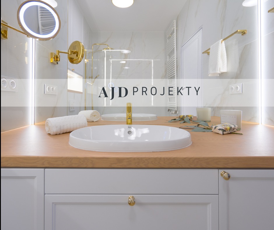 Jasna łazienka z białymi szafkami, drewnianym blatem, owalną umywalką, złotą armaturą i okrągłym lustrem z podświetleniem LED. Na blacie dekoracje: świeca, mydło i gałązki eukaliptusa.