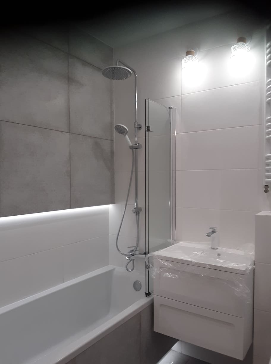 Nowoczesna łazienka z szarymi płytkami imitującymi beton, białą wanną z parawanem nawannowym, białą szafką pod umywalką jeszcze w folii ochronnej i zestawem prysznicowym z deszczownicą.