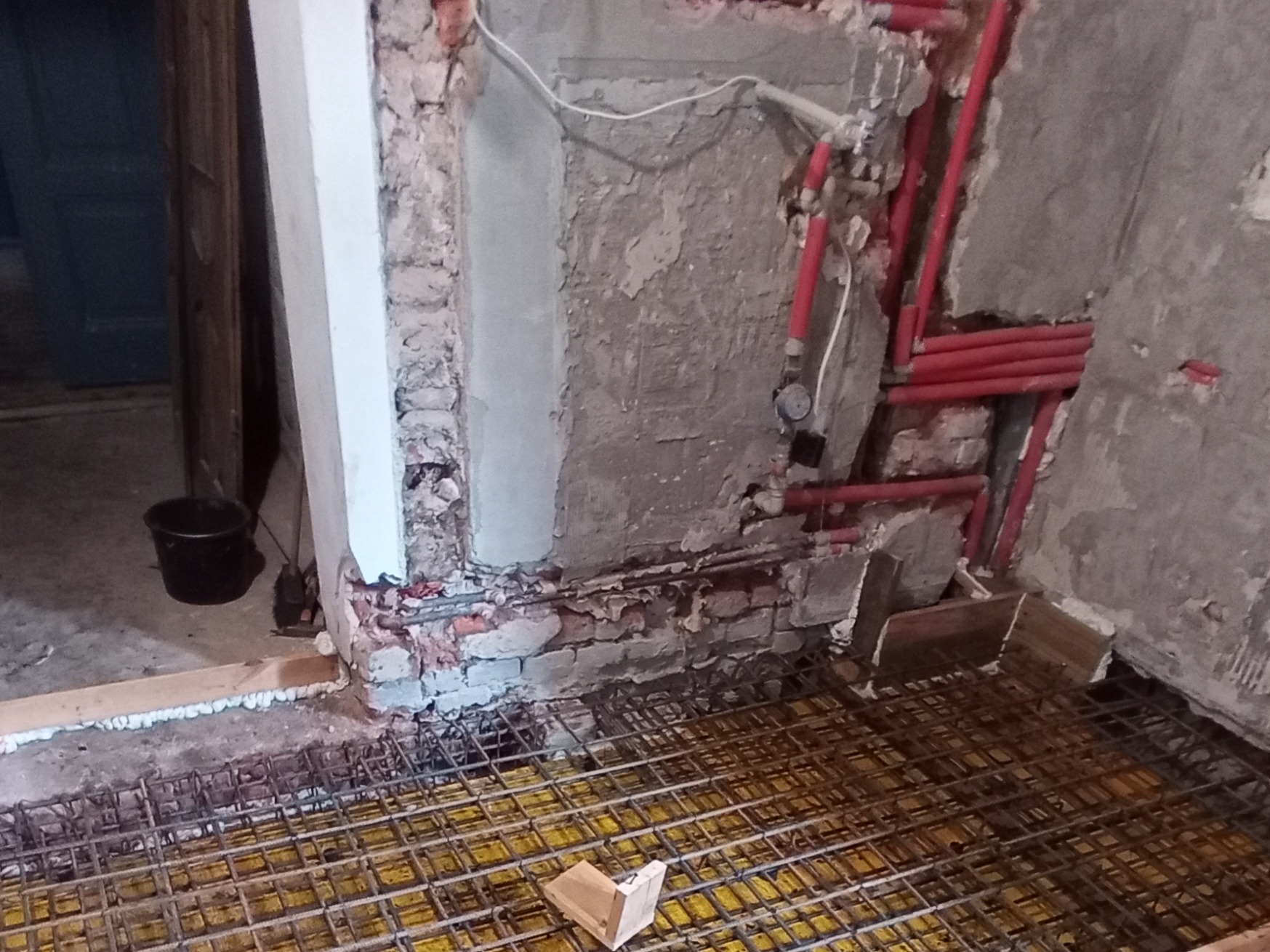 Przygotowanie podłoża pod ogrzewanie podłogowe w trakcie remontu, widoczne czerwone rury instalacji grzewczej oraz siatka zbrojeniowa na żółtej izolacji, ściany w stanie surowym.