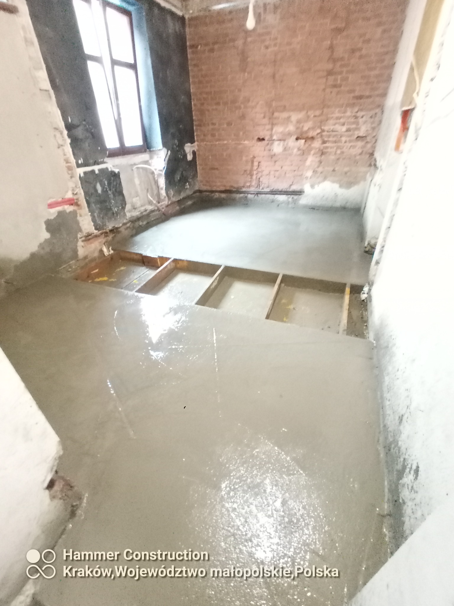 Wylewka samopoziomująca na podłodze w trakcie prac remontowych, widoczne drewniane ramy w posadzce, ściany w stanie surowym z widoczną cegłą i fragmentem okna.