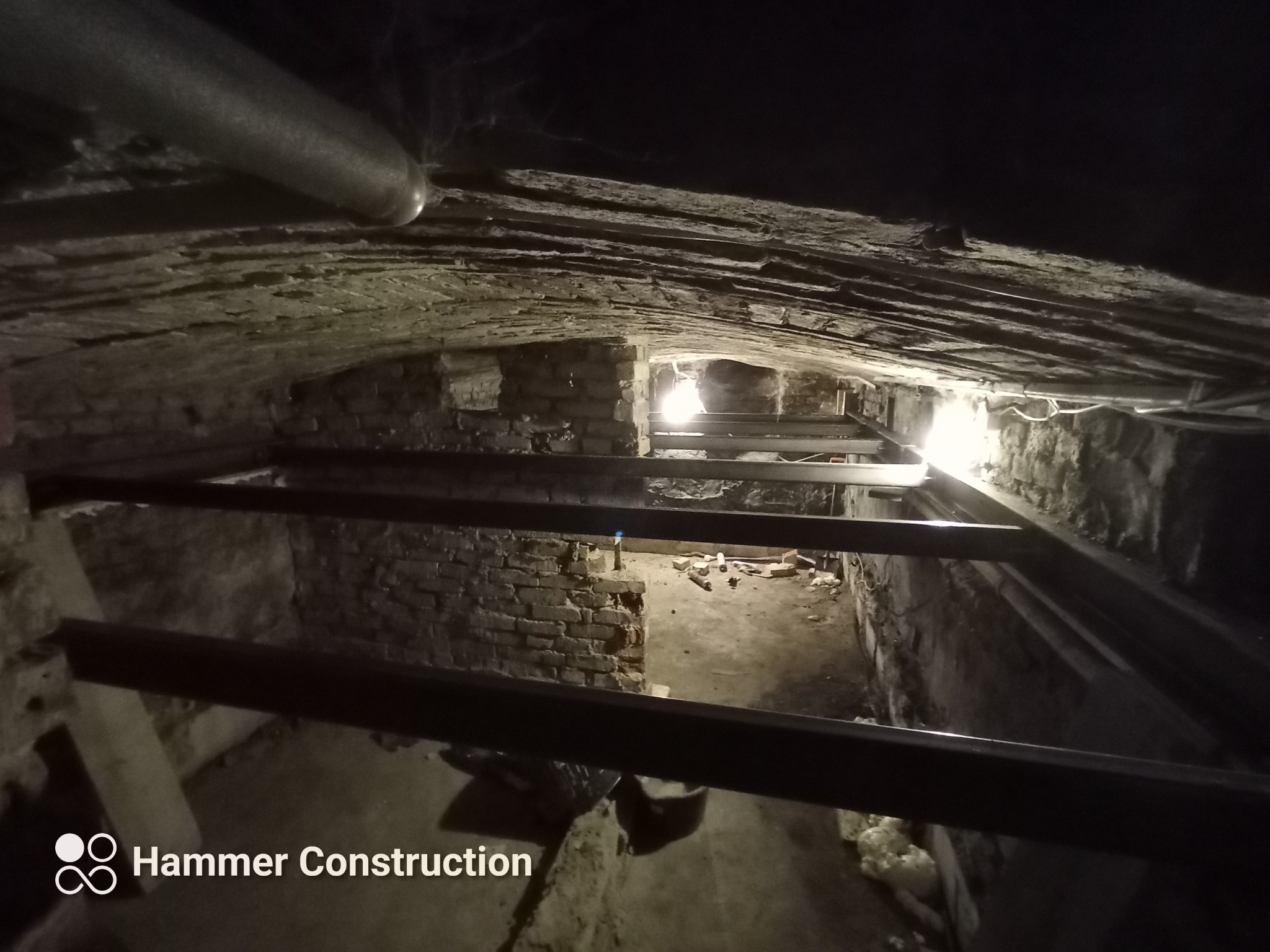 Wnętrze piwnicy z ceglanym sklepieniem łukowym, wzmocnione metalowymi belkami, oświetlone punktowo