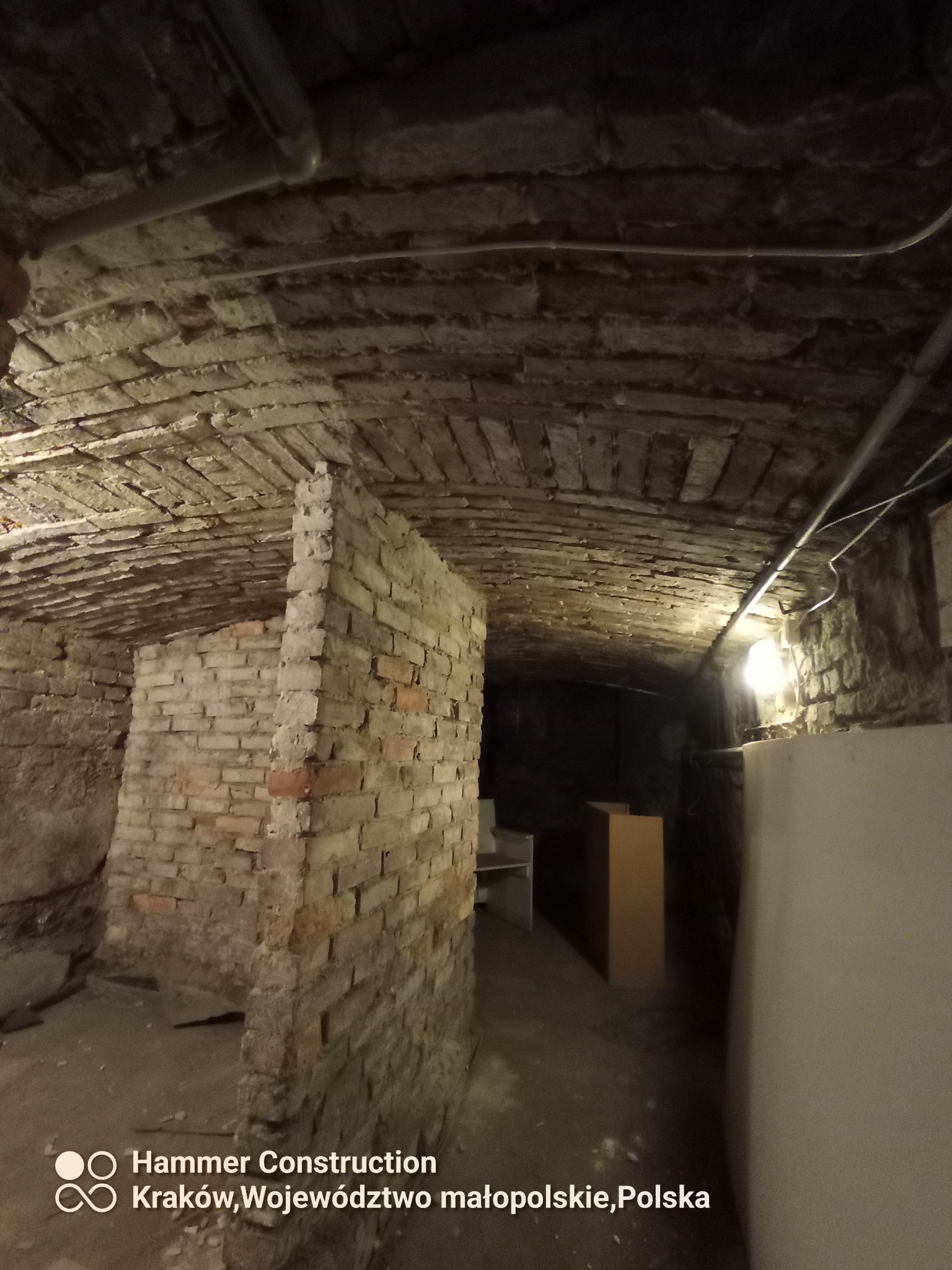 Krypta z ceglanym sklepieniem łukowym, fragment ściany działowej z cegieł, widoczne rury instalacyjne pod sufitem, słabe oświetlenie w głębi pomieszczenia.