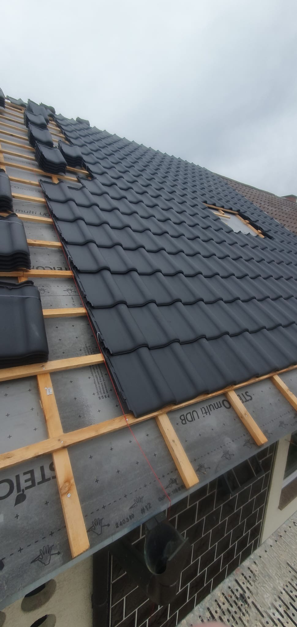 Częściowo pokryty czarną dachówką dach z widoczną konstrukcją drewnianą i membraną dachową, fragment dachu sąsiedniego budynku w tle.