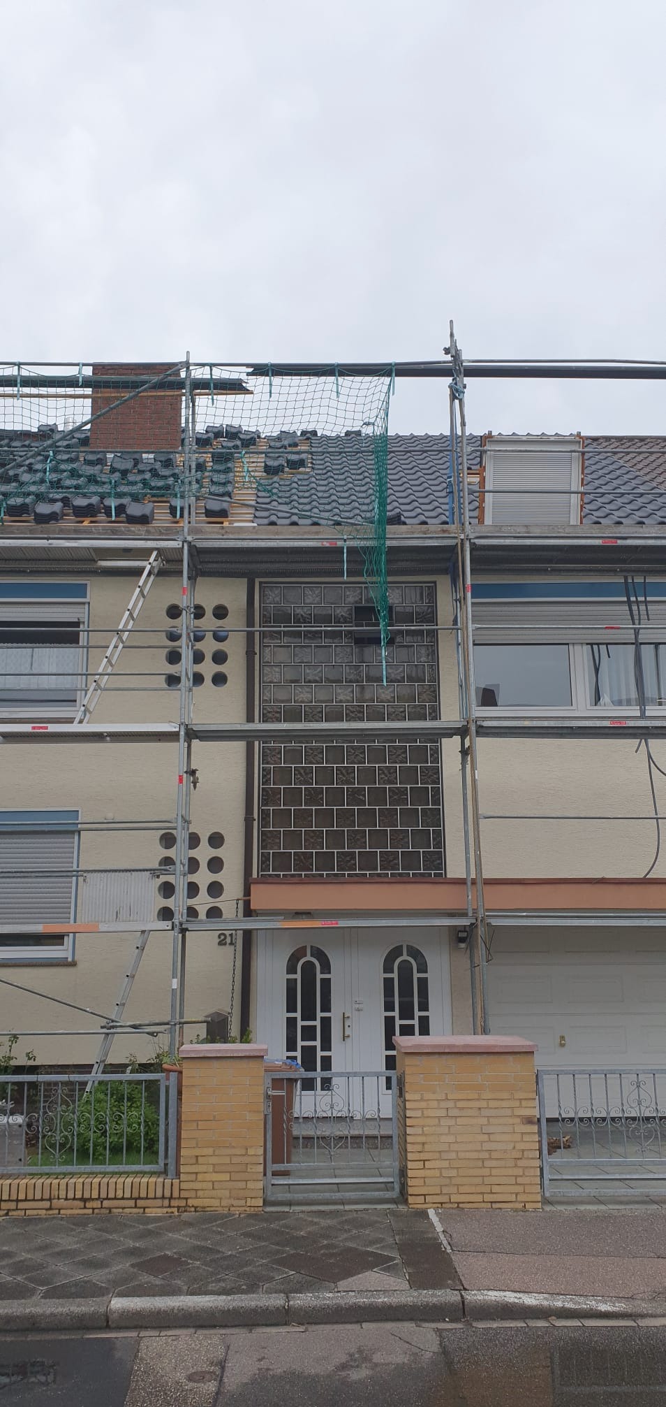 Dom jednorodzinny w trakcie wymiany pokrycia dachowego, widoczne rusztowania, częściowo położone nowe dachówki w kolorze grafitowym oraz komin z cegły.