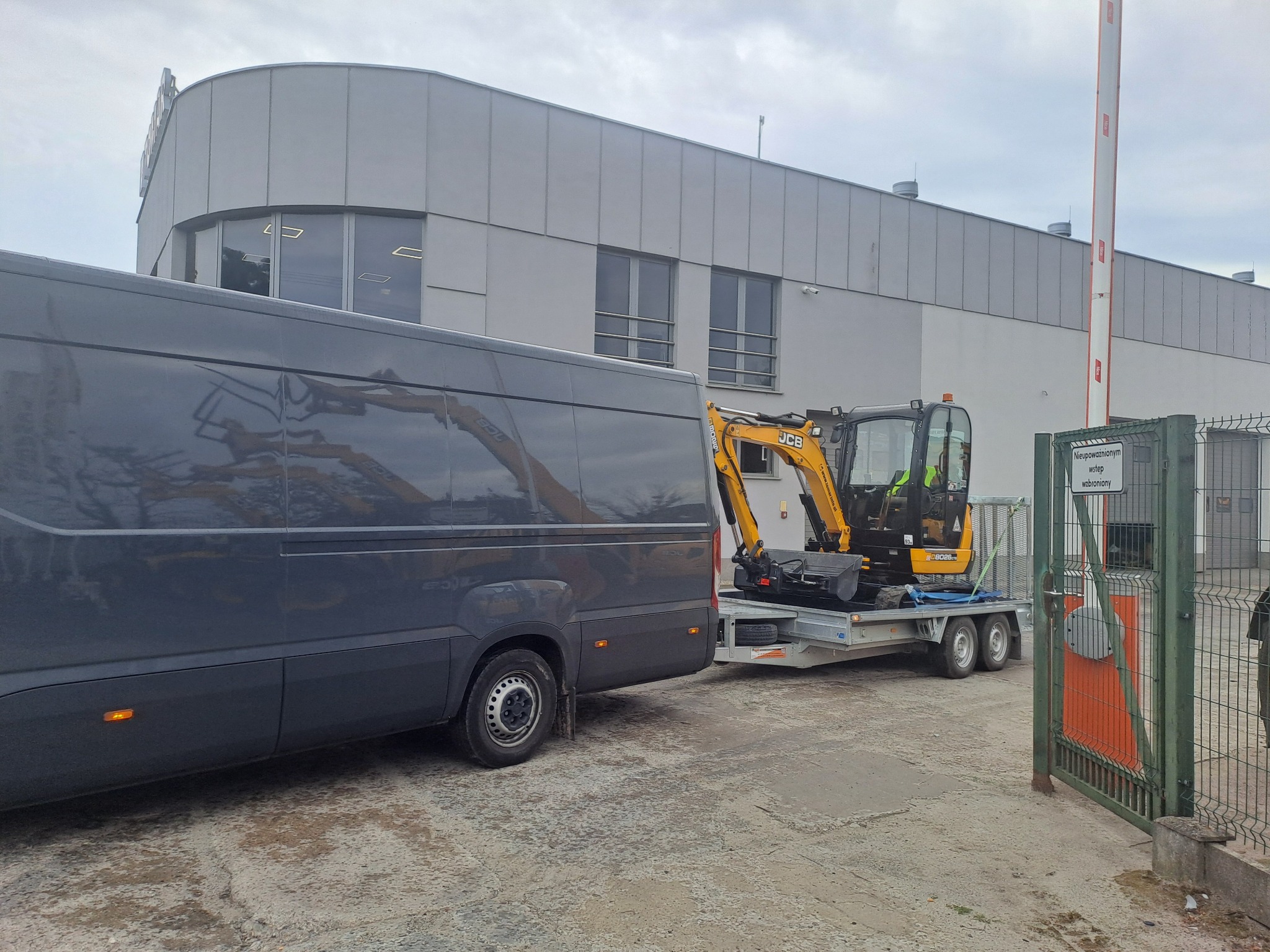 Żółta mini koparka JCB na lawecie, ciągnięta przez szary bus, przed budynkiem z szarej blachy trapezowej i zieloną bramą wjazdową.