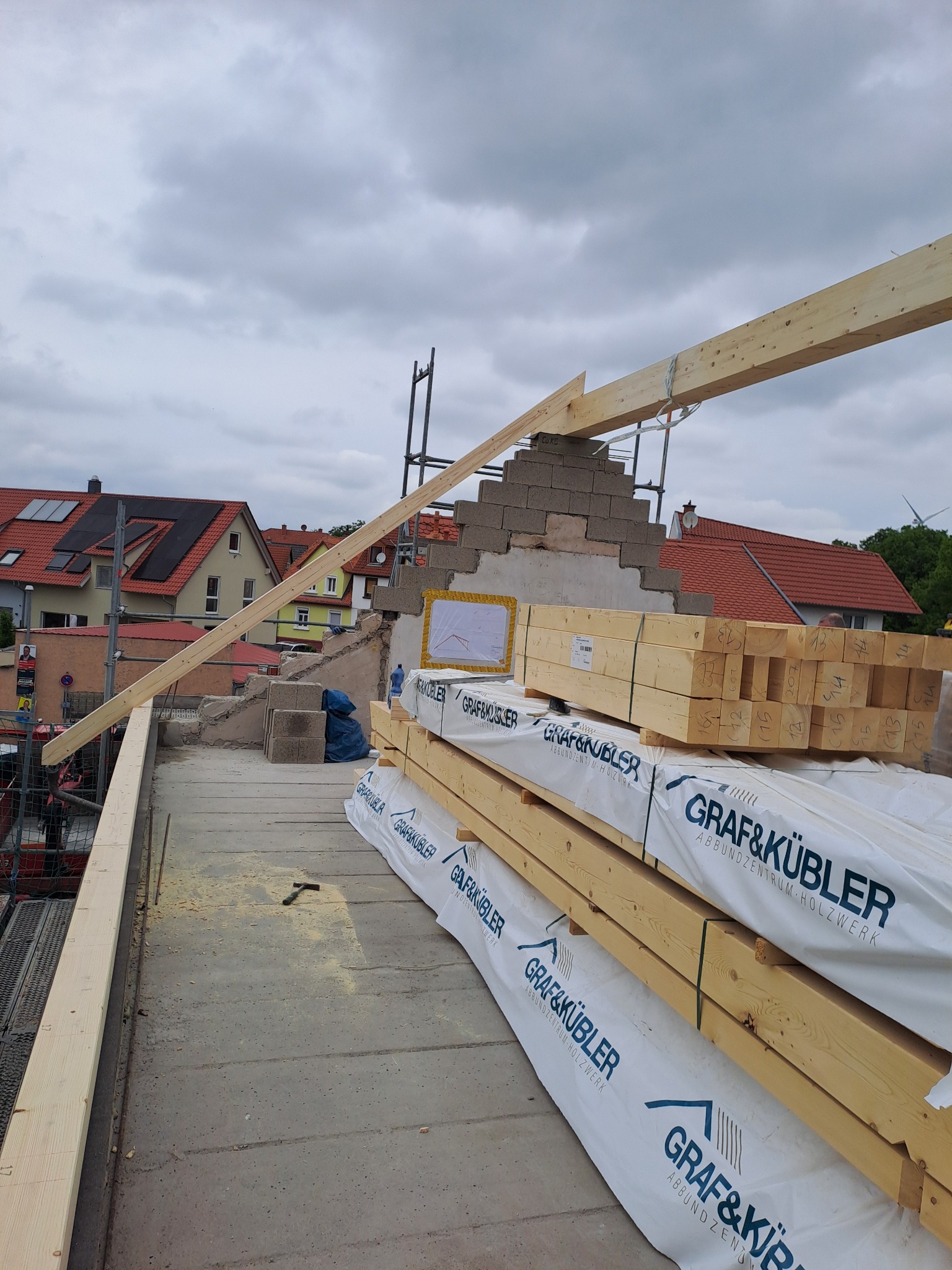 Konstrukcja dachu w trakcie budowy, widoczne elementy więźby dachowej, bloczki betonowe, drewniane belki, młotek i materiały budowlane z logo firmy Graf&Kübler na tle zabudowy miejskiej...