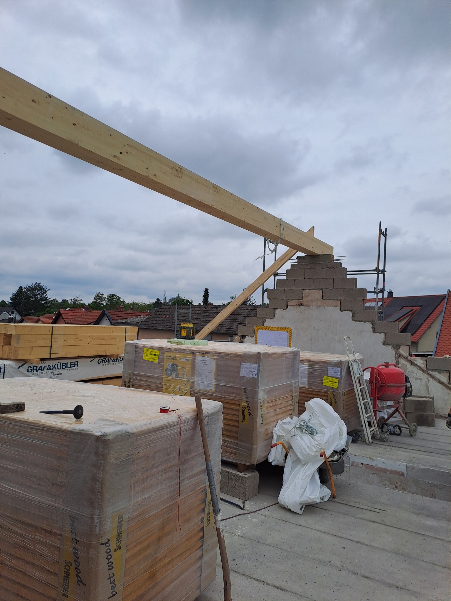 Budowa dachu: widok na palety z drewnem, betonowe bloczki, podnośnik, mikser i fragment konstrukcji dachu pod pochmurnym niebem.