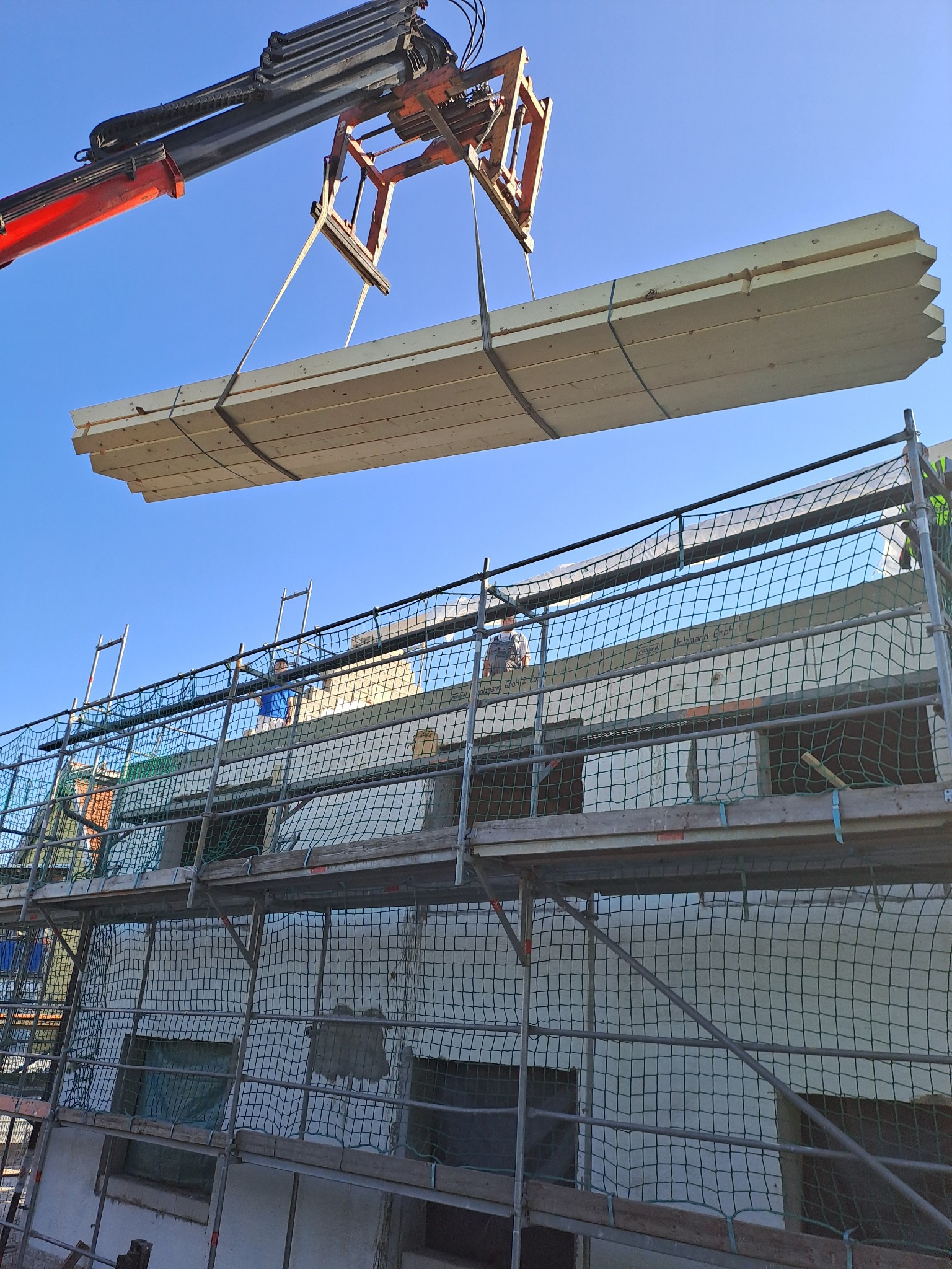 Ciężki dźwig podnosi pakiet drewnianych belek na placu budowy, widoczne rusztowania, pracownicy i ściany budynku w surowym stanie.
