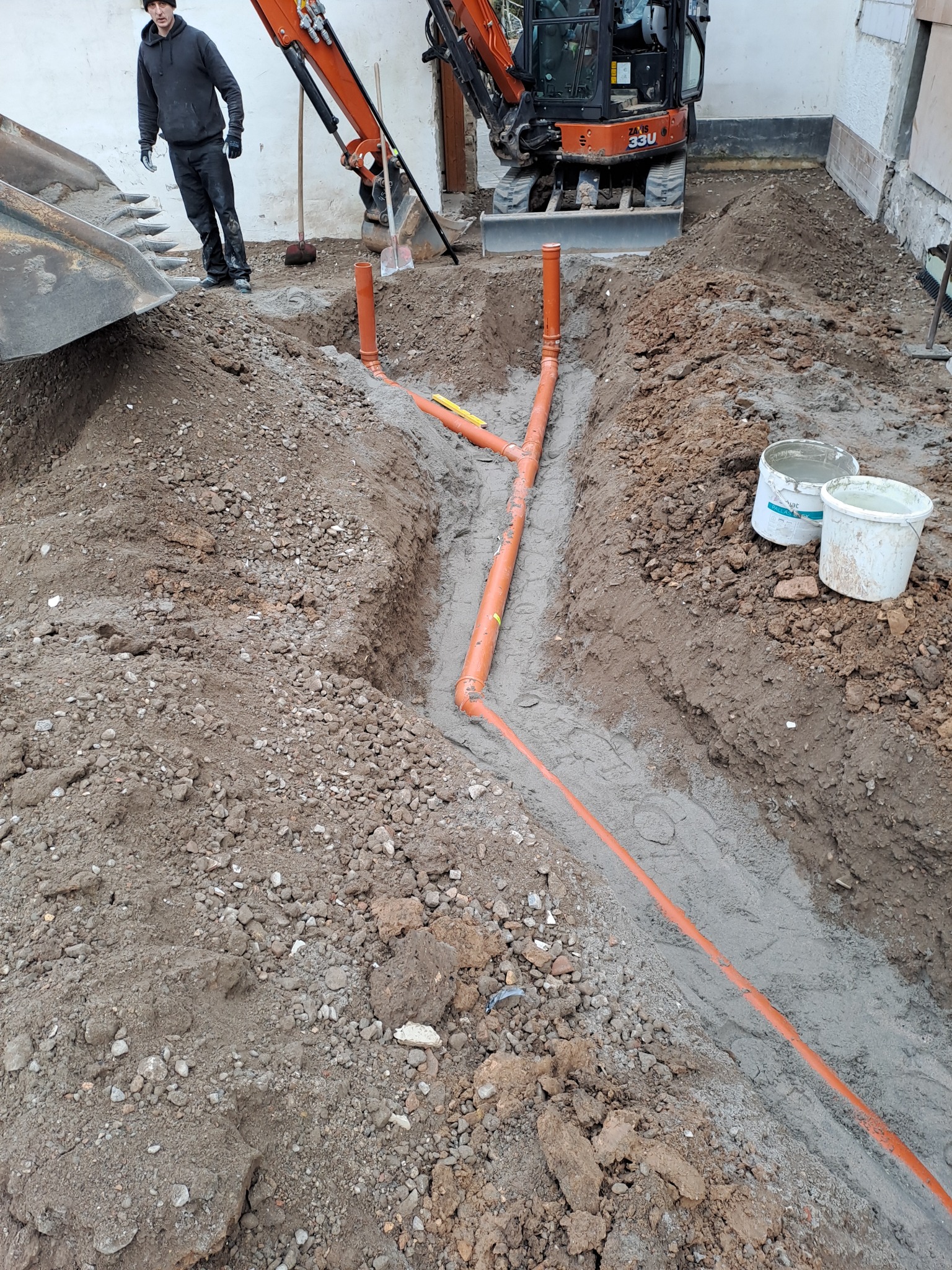Wykop z ułożonymi pomarańczowymi rurami kanalizacyjnymi, częściowo zasypany piaskiem, widoczny mini-koparka i pracownik w tle.