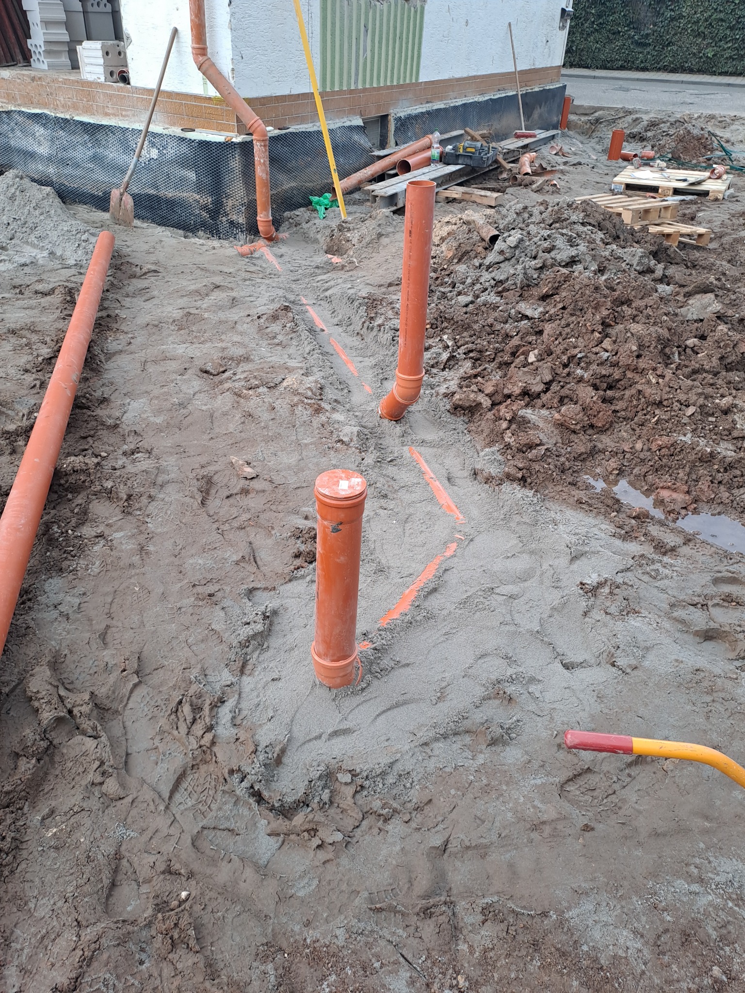 Instalacja pomarańczowych rur kanalizacyjnych w wykopie, widoczne ślady prac ziemnych, piasek, narzędzia i materiały budowlane w tle.