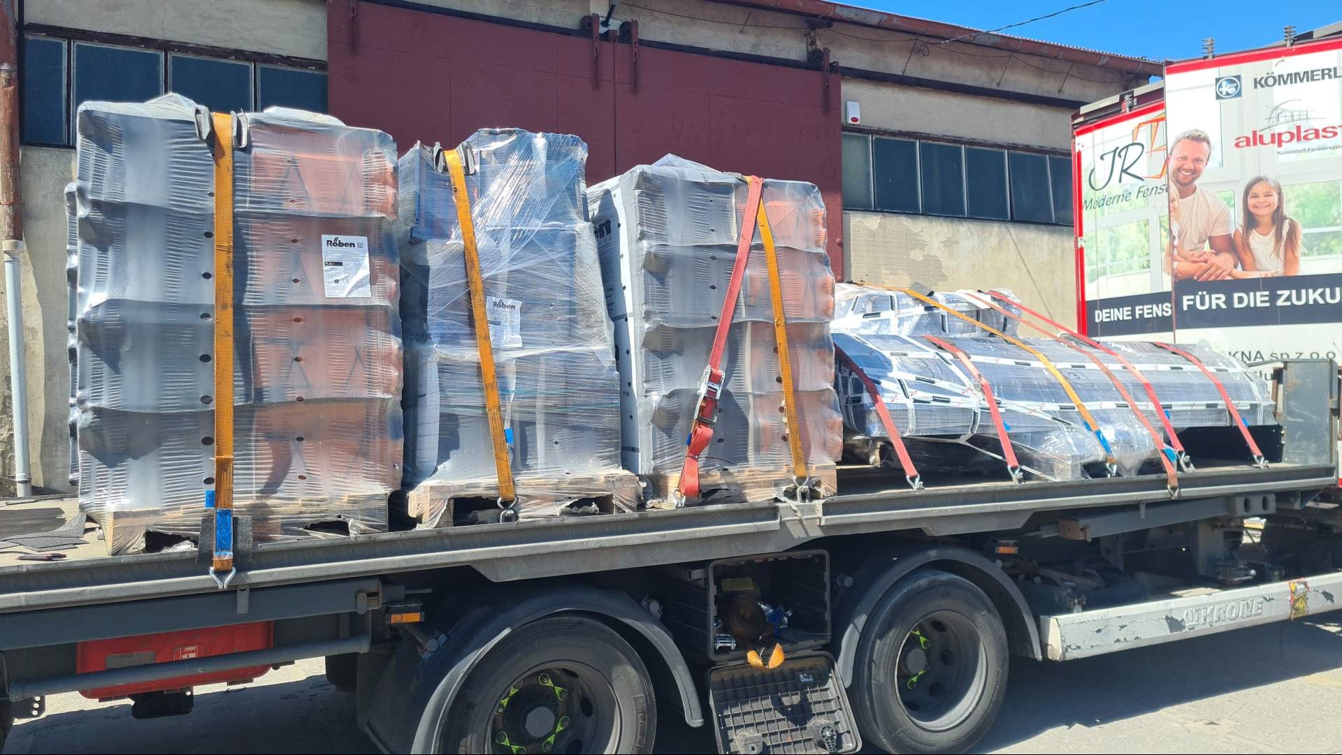 Ciężarówka z załadunkiem palet z dachówką ceramiczną Röben oraz palet z oknami aluminiowymi, zabezpieczonych pasami transportowymi, na tle budynku przemysłowego i reklamy okien.