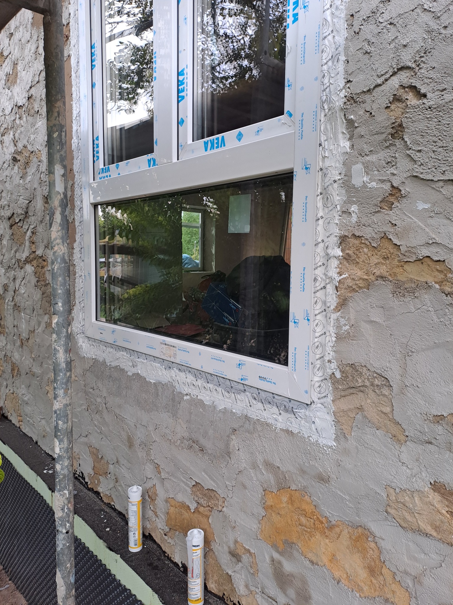 Montaż białego okna z PCV w surowej ścianie z widocznym ociepleniem i tubami uszczelniacza Wurth u podstawy. Okno z naklejkami VEKA i Hoppe, widoczny odblask drzew.