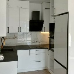 Komodus - Biała, lakierowana zabudowa kuchenna z czarnymi, podłużnymi uchwytami, czarnym okapem i blatem, oraz marmurowym panelem nad blatem, oświetlona listwą LED.