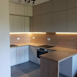 Komodus - Nowoczesna kuchnia w kształcie litery U z białymi i szarymi frontami, drewnianym blatem i podświetleniem LED pod szafkami wiszącymi oraz czarną lampą sufitową. Widoczny fragment podłogi z szarych...