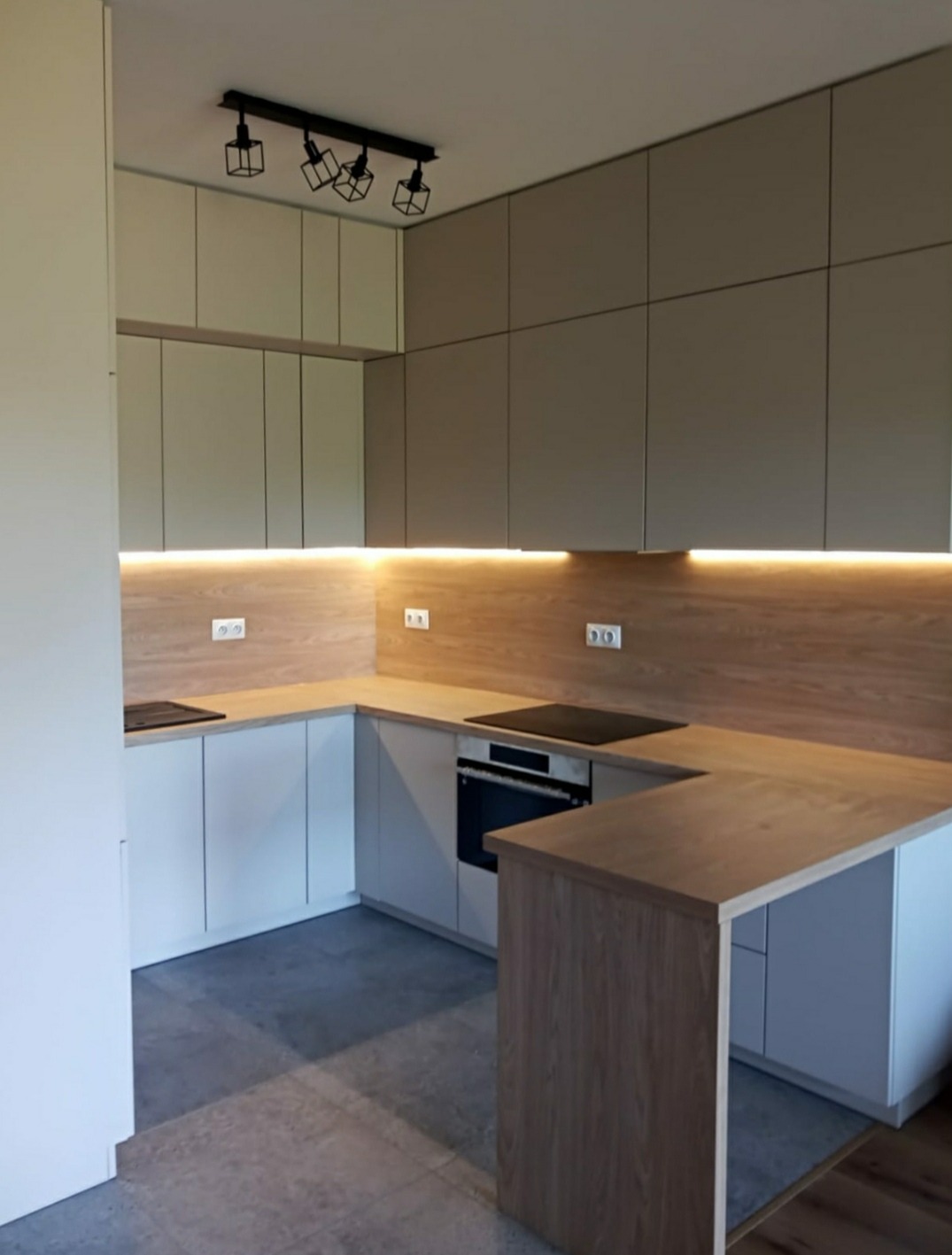 Nowoczesna kuchnia w kształcie litery U z białymi i szarymi frontami, drewnianym blatem i podświetleniem LED pod szafkami wiszącymi oraz czarną lampą sufitową. Widoczny fragment podłogi z szarych...