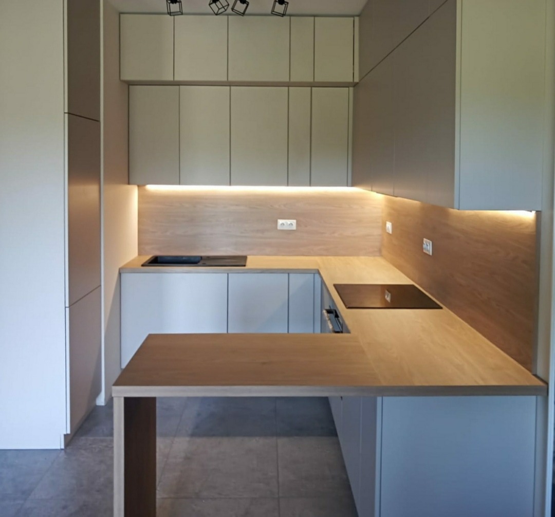 Nowoczesna, jasna kuchnia z drewnianym blatem i oświetleniem LED pod szafkami, widoczny fragment wyspy kuchennej.