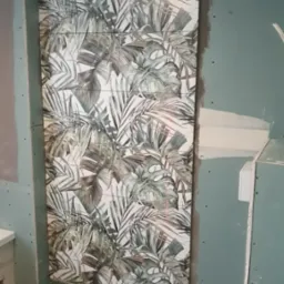 Fragment ściany w trakcie montażu płytek z motywem tropikalnych liści, widoczne laserowe linie poziomowania, surowy stan wykończenia pomieszczenia.