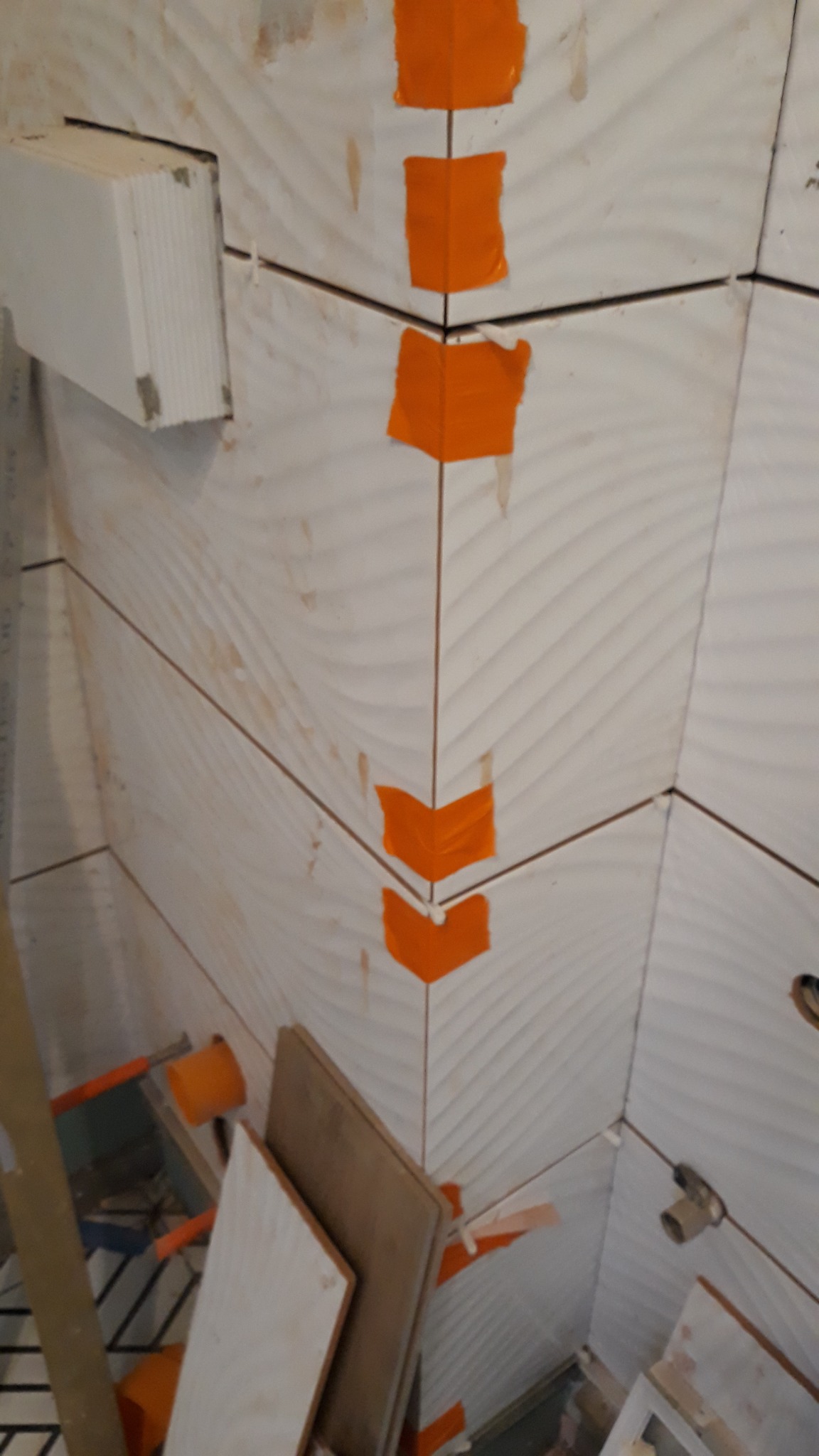 Montaż płytek ściennych w łazience z użyciem pomarańczowej taśmy maskującej, widoczne narzędzia i materiały budowlane w tle.