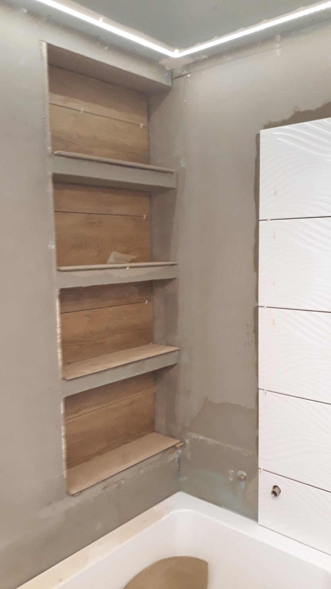 Niewykończona wnęka z drewnopodobnymi płytkami i półkami w łazience, białe płytki ułożone pionowo na ścianie, widoczna wanna, surowy tynk, oświetlenie LED w suficie.