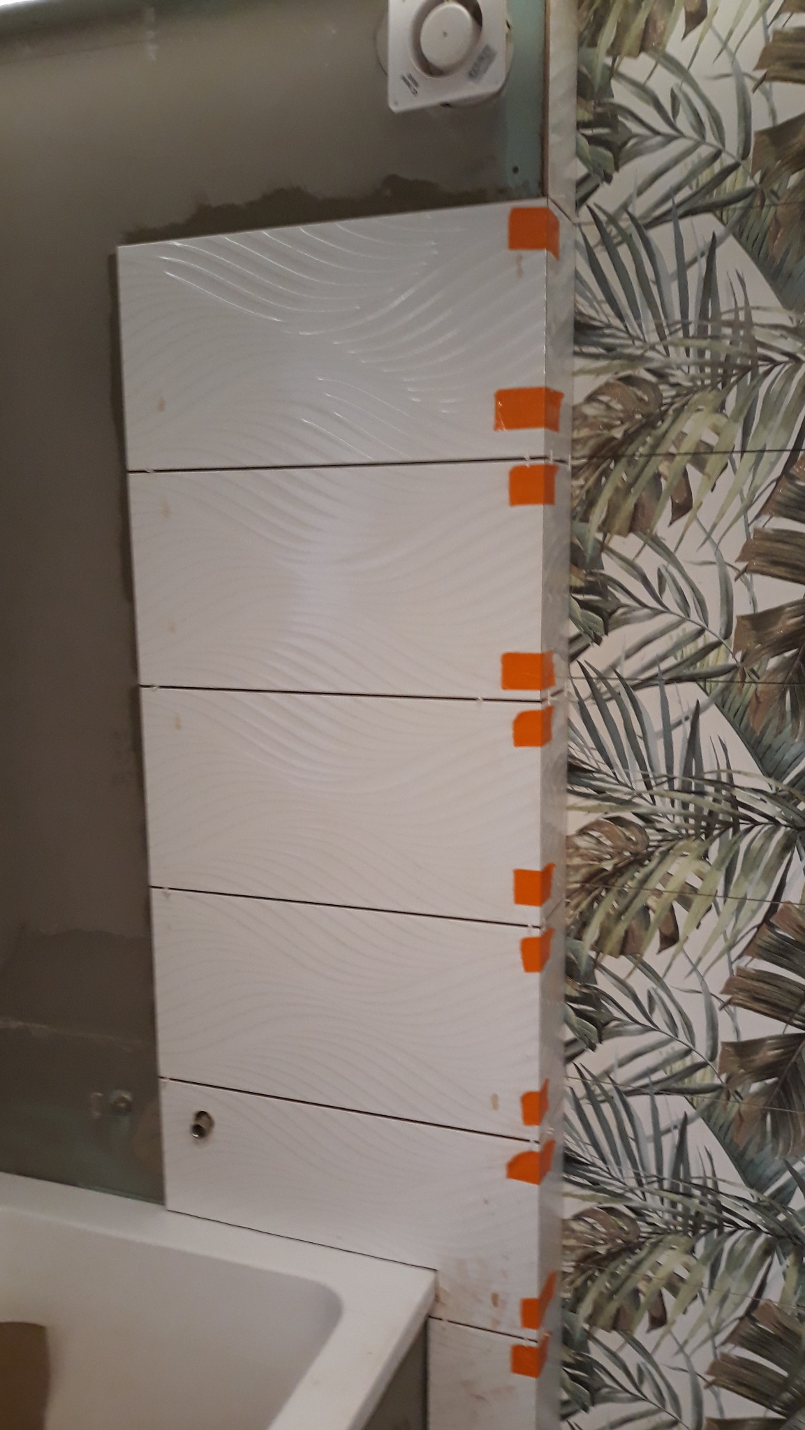 Układanie białych, poziomych płytek z delikatnym wzorem fal, zabezpieczonych pomarańczowymi krzyżykami dystansowymi, na ścianie w łazience z widoczną wanną i tapetą w tropikalne liście.