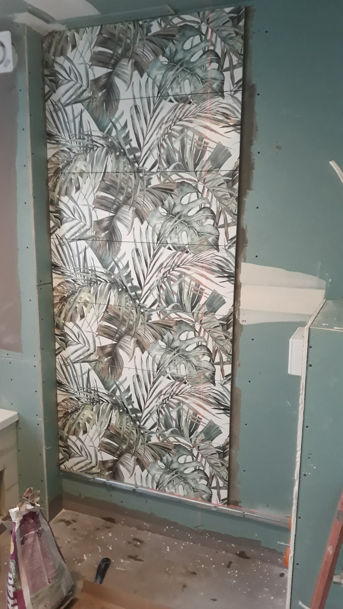 Fragment ściany w trakcie montażu płytek z motywem tropikalnych liści, widoczne laserowe linie poziomowania, surowy stan wykończenia pomieszczenia.