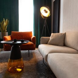 Tuwima 48 Sp. z o.o. - Stylowe wnętrze z sofą w odcieniach beżu, pomarańczowym fotelem i designerskim stolikiem kawowym z dwoma filiżankami na dywanie w nowoczesnym biurze.