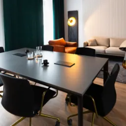 Nowoczesna sala konferencyjna z dużym stołem, czarnymi krzesłami na złotych nogach, zielonymi zasłonami i stylowym oświetleniem. Widoczny również sofa i stolik.