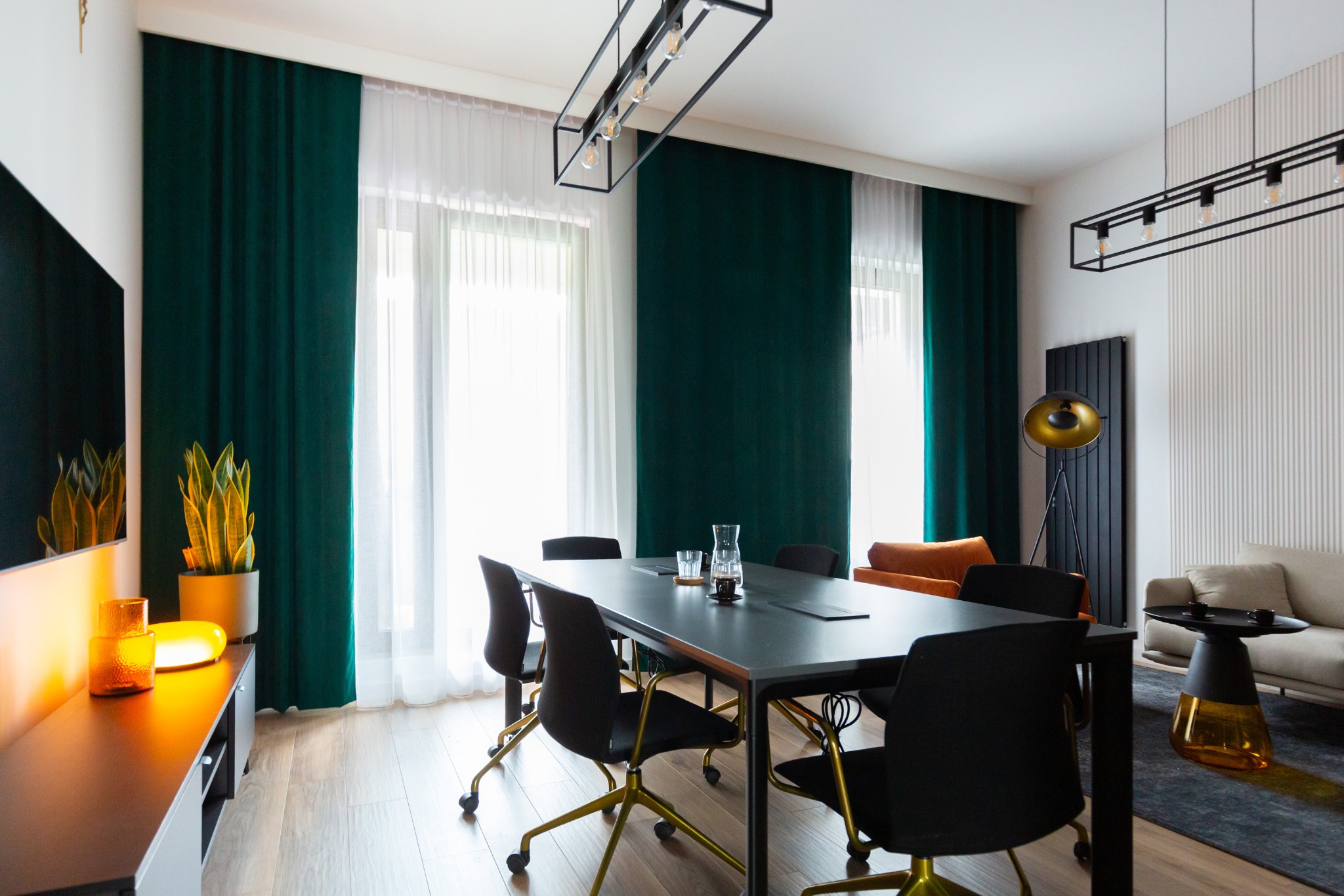 Nowoczesna sala konferencyjna z dużym stołem, czarnymi krzesłami na złotych nogach i zielonymi zasłonami. Jasne wnętrze z elementami dekoracyjnymi.