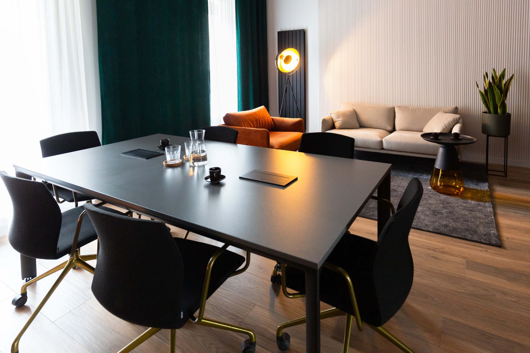 Nowoczesna sala konferencyjna z dużym stołem, czarnymi krzesłami na złotych nogach, zielonymi zasłonami i stylowym oświetleniem. Widoczny również sofa i stolik.
