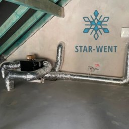 Star-Went PATRYK DULĘBA - Zamontowany system rekuperacji z czarną centralą i srebrnymi, elastycznymi przewodami w pomieszczeniu na poddaszu z widoczną konstrukcją dachu i logo firmy STAR-WENT na ścianie.