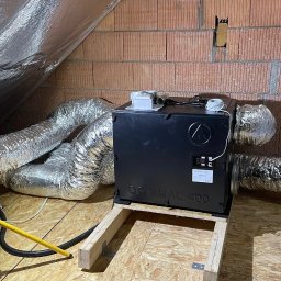 Star-Went PATRYK DULĘBA - Czarna jednostka rekuperacyjna Optimal 400 zamontowana na drewnianej ramie w surowym pomieszczeniu na poddaszu z widocznymi przewodami wentylacyjnymi i izolacją folią aluminiową.