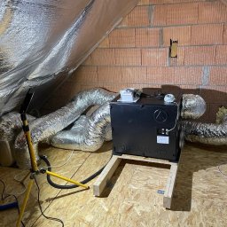 Star-Went PATRYK DULĘBA - Czarna centrala wentylacyjna Optimal 400, zamontowana na drewnianej konstrukcji na poddaszu, z podłączonymi elastycznymi przewodami wentylacyjnymi, widoczna lampa robocza na trójnogu.