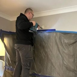 Star-Went PATRYK DULĘBA - Pracownik na drabinie wierci otwór w ścianie, zabezpieczonej folią i taśmą malarską, używając wiertarki akumulatorowej Makita.