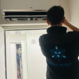Star-Went PATRYK DULĘBA - Instalator w firmowej bluzie STAR-WENT montuje białą klimatyzację LG na ścianie w pomieszczeniu z szafą elektryczną. Widoczne przewody w szafie.