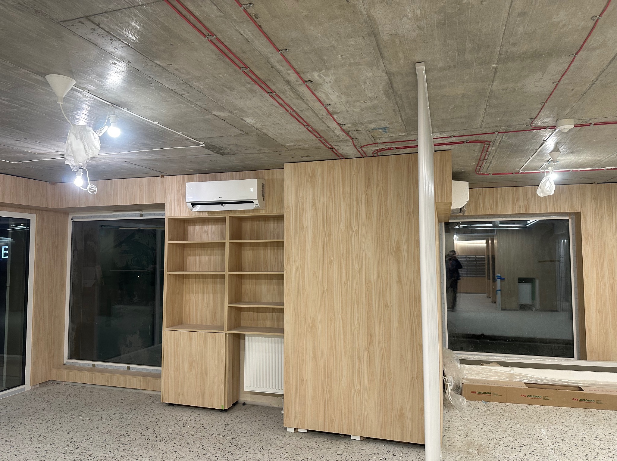 Wnętrze w trakcie prac wykończeniowych: zamontowana klimatyzacja LG na ścianie, drewniana zabudowa regałowa, widoczne instalacje elektryczne pod betonowym sufitem, częściowo zasłonięte oświetlenie,...