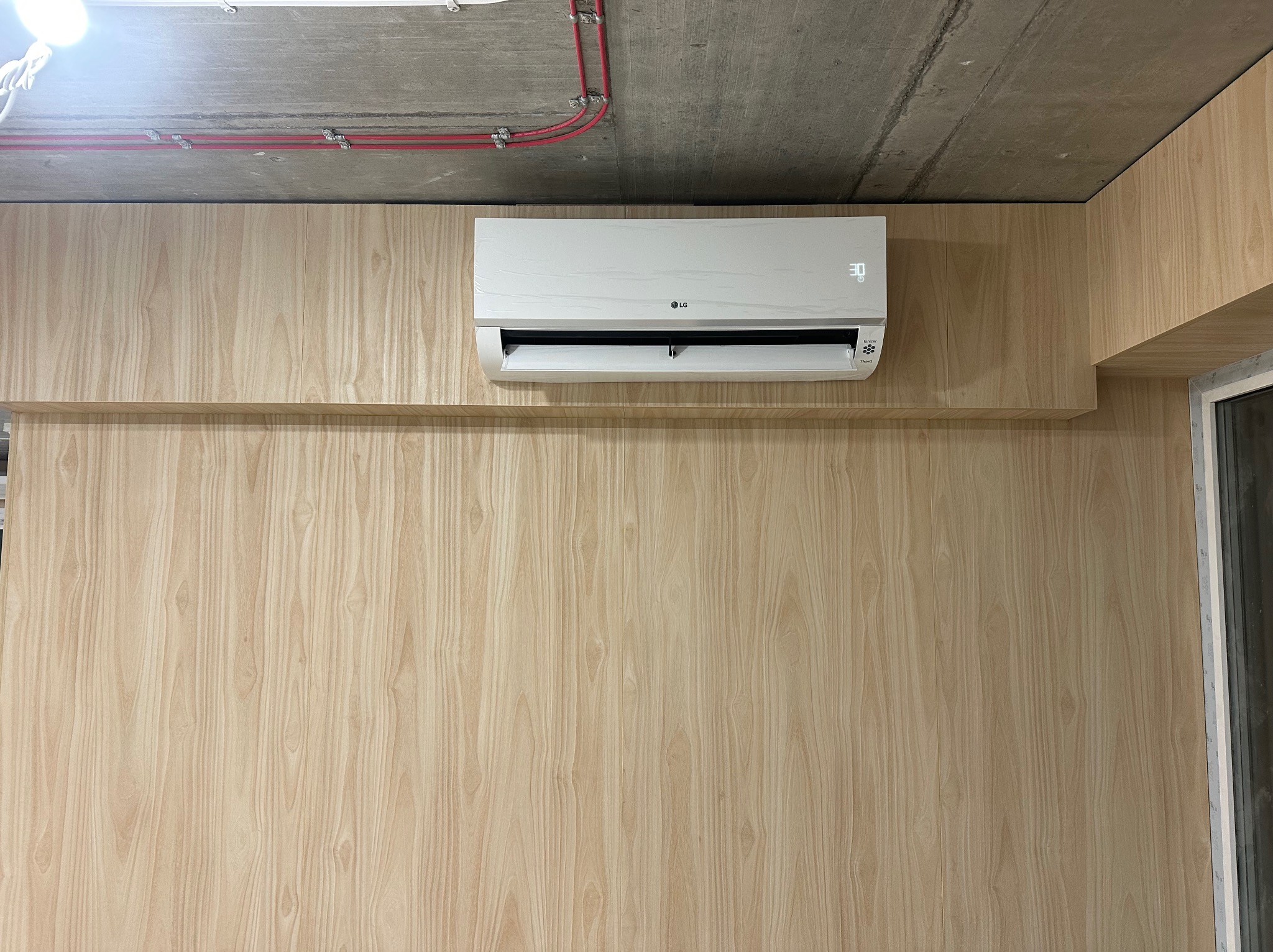 Zamontowany, biały klimatyzator LG na ścianie wykończonej panelami imitującymi jasne drewno, widoczne przewody elektryczne pod betonowym sufitem.
