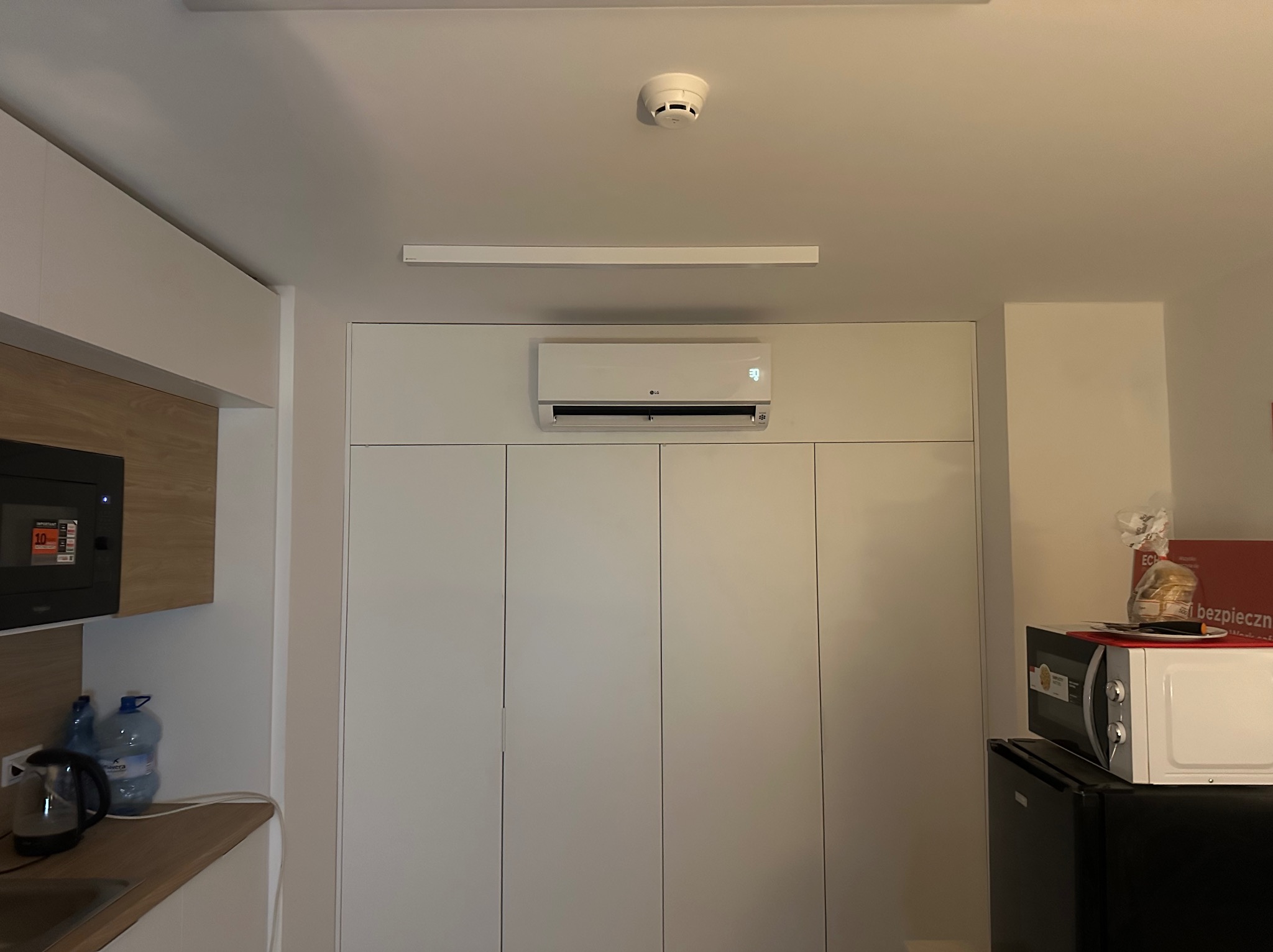 Biały klimatyzator LG zamontowany nad szafą w zabudowie w jasnej kuchni, widoczny czujnik dymu na suficie i fragment blatu z czajnikiem.