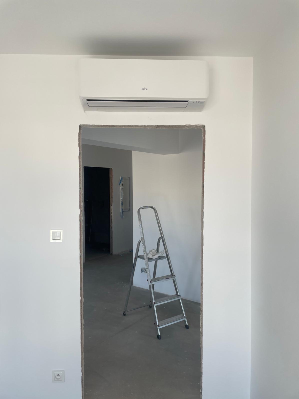 Biała jednostka klimatyzacyjna Fujitsu zamontowana nad otworem drzwiowym w surowym, białym wnętrzu, z rozstawioną drabiną aluminiową w tle.